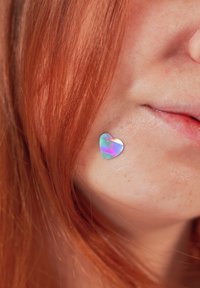 Personne aux cheveux roux avec un petit autocollant irisé en forme de cœur sur la joue droite près de la bouche.