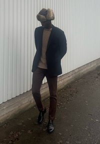 Homme portant un chapeau orné de fourrure, un pull rayé, un blazer sombre, un pantalon marron et des chaussures noires, marchant le long d'un mur ondulé sur un chemin humide.