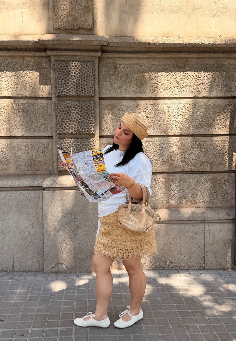 Femme en tenue d'été, portant un chapeau et tenant un sac tressé, lit une carte en se tenant sur un trottoir ensoleillé de la ville près d'un mur en pierre.