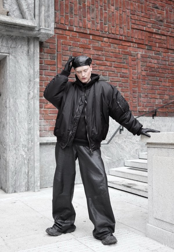 Persoană purtând beret negru din piele, mănuși, geacă bomber supradimensionată, pantaloni largi din piele, stând lângă un parapet de piatră, în fața unui perete de cărămidă.