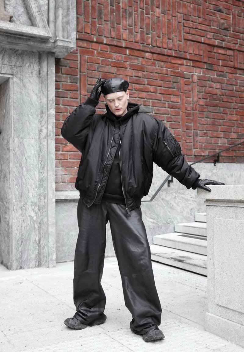 Personne portant un béret en cuir noir, des gants, une veste bomber oversize, un pantalon large en cuir, debout près d'une balustrade en pierre contre un mur de briques.