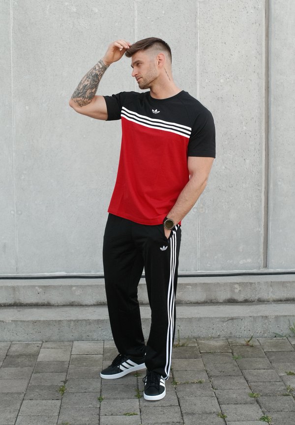 Muž s tetovanou paží v černém a červeném outfitu Adidas stojí na dlážděné zemí u betonové zdi a dotýká se vlasů.