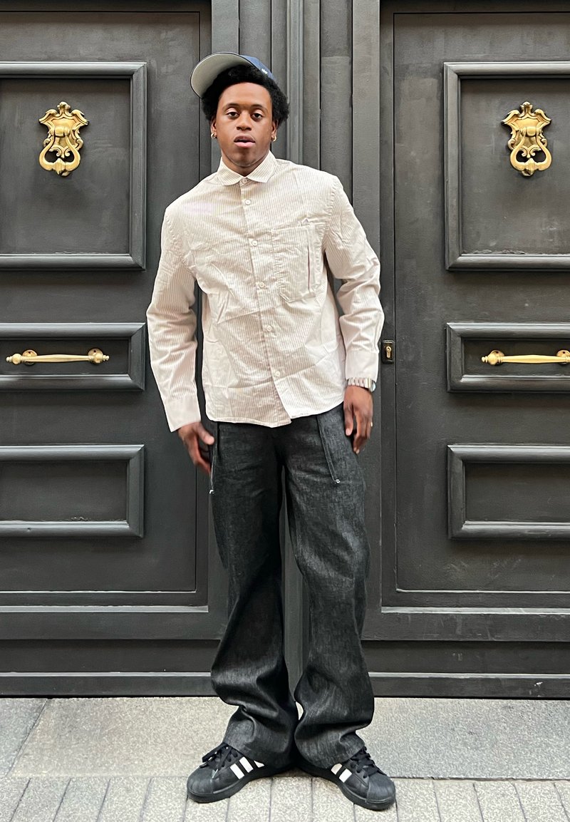 Chemise à rayures blanches à boutons avec des manches longues, pantalon en denim noir amp; large, et baskets noires avec des rayures blanches ; casquette portée sur le côté.