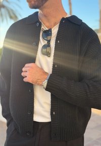 Άνδρας με μαύρη πλεκτή ζακέτα, λευκό μπλουζάκι και ρολόι κρατά τη ζακέτα με γυαλιά ηλίου που κρέμονται στο μπλουζάκι, σε εξωτερικό χώρο με φόντο φοίνικα.