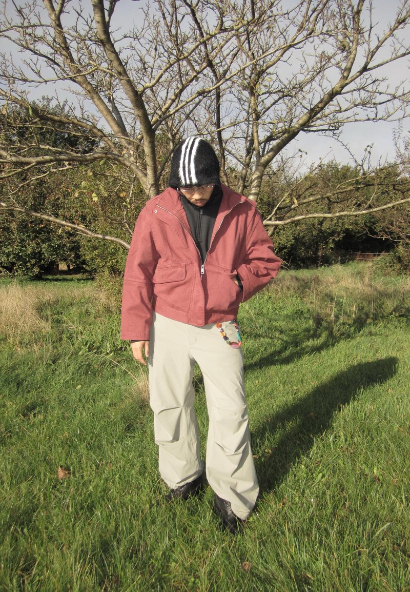 Red zip-up jacket, black hoodie, beige cargo pants, black boots; striped beanie; standing on grass with trees in background.