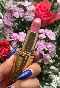 Hand die een roze L'Oréal lipstick vasthoudt met een glanzende gouden huls, blauwe glinsterende nagels, en roze en rode bloemen op de vervaagde achtergrond.