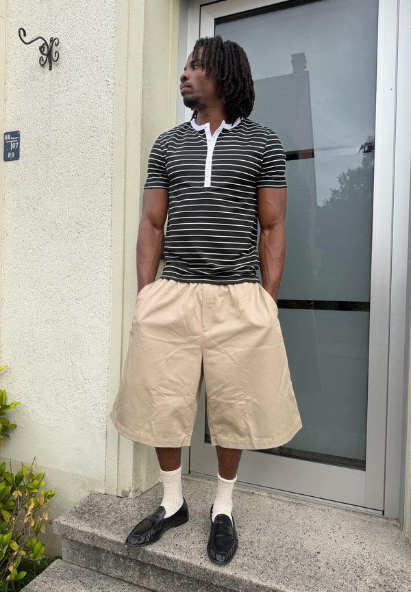 Uomo con dreadlocks che indossa una camicia a righe bianche e nere, pantaloncini beige largi, calzini bianchi e mocassini neri, in piedi su gradini di cemento vicino a una porta.