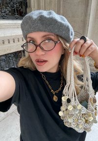 Világosszürke makramé táska fényvisszaverő arany kör alakú díszekkel, egy személy kezében, aki fekete pólót és szürke sapkát visel. Átlátszó keretes szemüveg.
