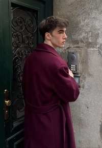 Manteau bordeaux avec ceinture, debout à côté d'un mur gris texturé et d'un interphone, devant une porte verte ornée de ferronnerie.