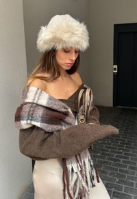 Knitted white hat with faux fur trim, brown oversized sweater, multi-coloured fringed scarf, and cream satin skirt in an urban setting.
