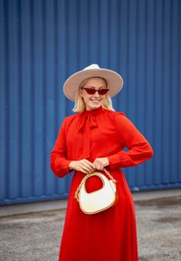 Červené šaty s dlhým rukávom a detailom mašle, doplnené krémovou a hnedou kabelkou. Pre veľkú svetlo šedú čiapku a červené slnečné okuliare dokončujú vzhľad.