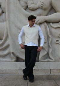 Off-white brezrokavnik, složen čez belo srajco, v kombinaciji s črnimi hlačami s črtami. Gladek material, minimalističen dizajn, obut v temne čevlje.