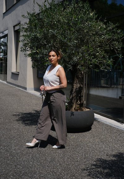 Blusa sin mangas de color claro combinada con pantalones grises holgados y zapatos de tacón alto blancos. De pie junto a un olivo en un entorno moderno.