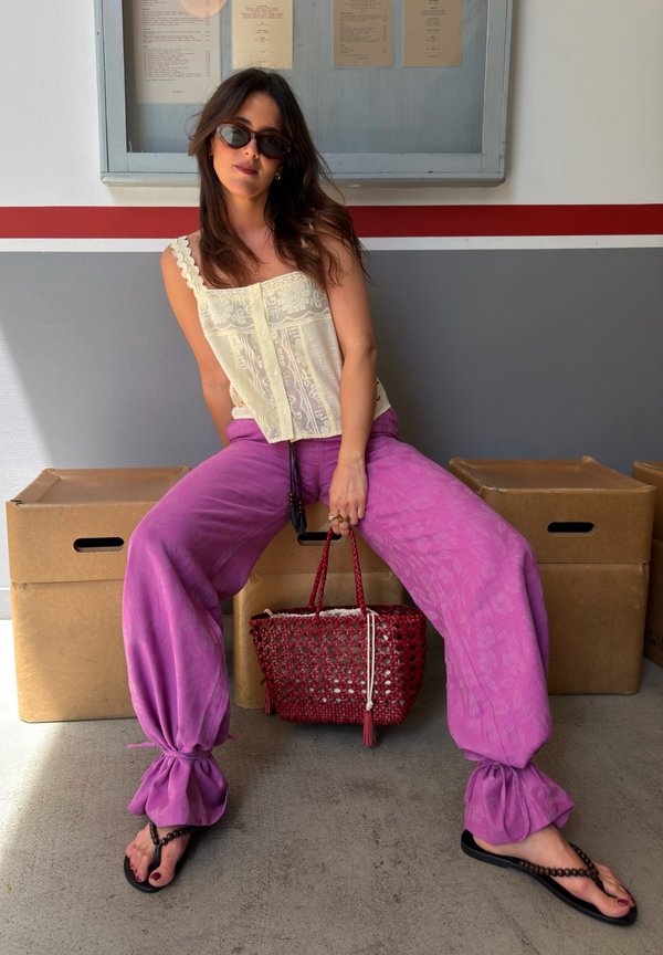 Mujer con el pelo largo y gafas de sol sentada sobre una caja, lleva un top de encaje color crema, pantalones morados sueltos, chanclas y sostiene un bolso rojo de mimbre.