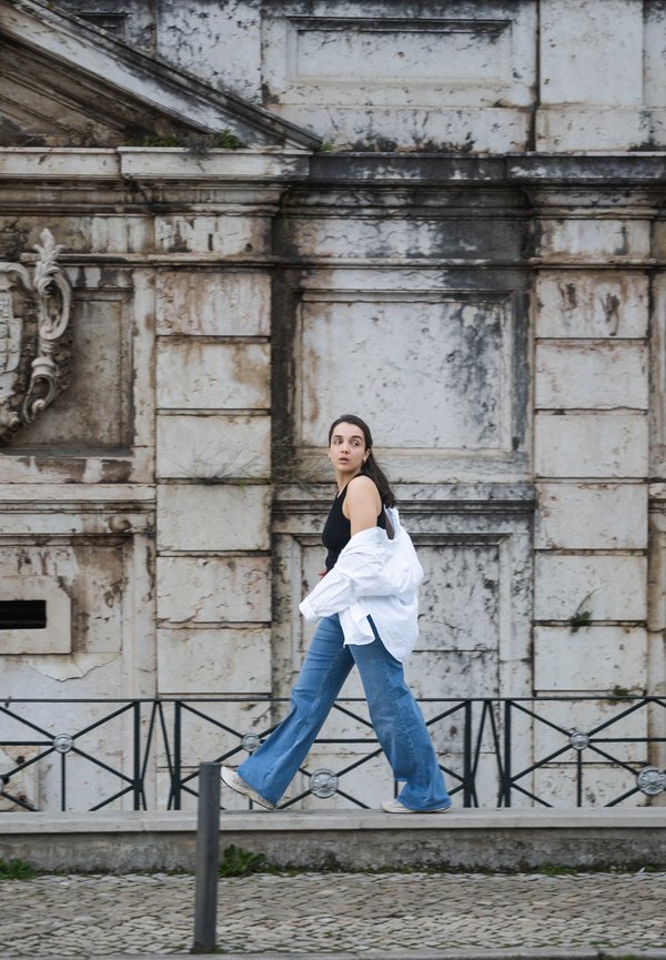 Una persona cammina lungo un sentiero di pietra indossando una canotta nera, una camicia bianca oversize e dei jeans a zampa d'elefante bleu chiaro con delle sneakers bianche.