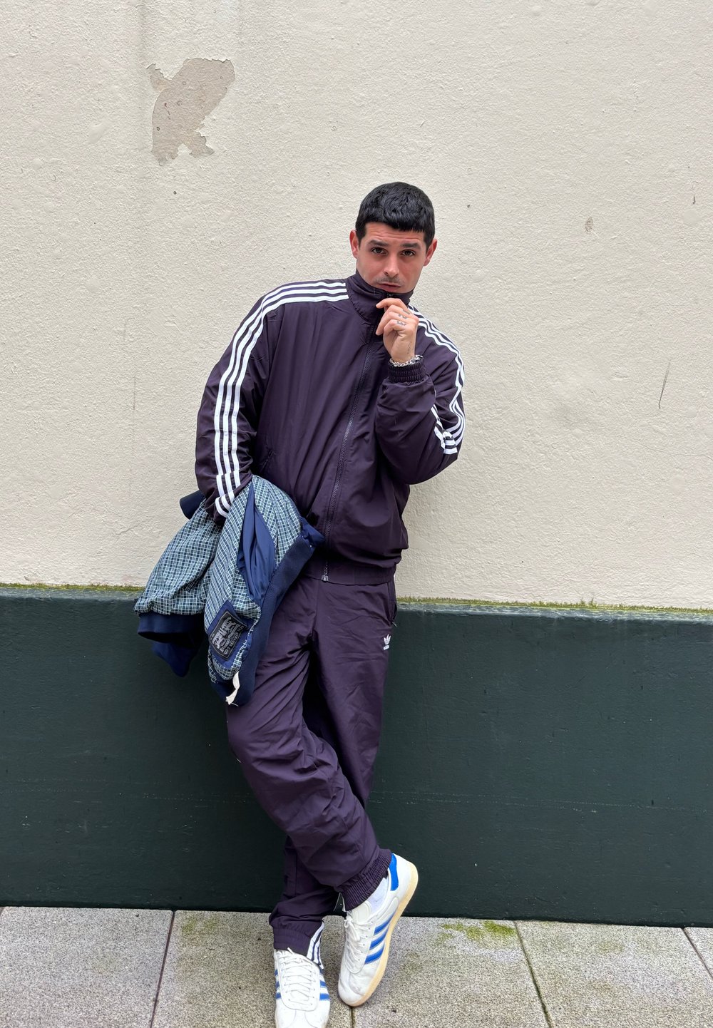 Survêtement violet avec des rayures blanches, associé à une veste en plaid bleu texturé. Debout contre un mur clair, chaussé de baskets blanches et bleues.