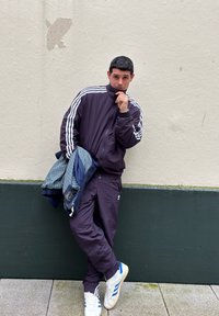Survêtement violet avec des rayures blanches, associé à une veste en plaid bleu texturé. Debout contre un mur clair, chaussé de baskets blanches et bleues.