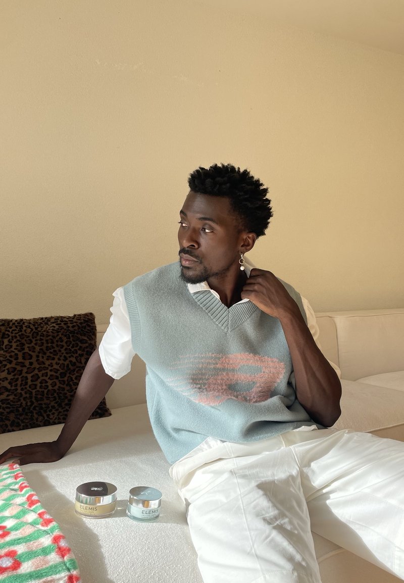 Gilet tricoté bleu clair avec un motif rose, porté sur une chemise blanche. Pantalon blanc, deux pots de cosmétiques étiquetés "ELEMIS", et un oreiller à imprimé léopard.