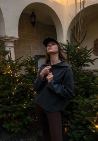 Femme portant une veste sombre et une casquette, debout parmi des sapins décorés de lumières dans une cour aux murs voûtés et une lanterne.
