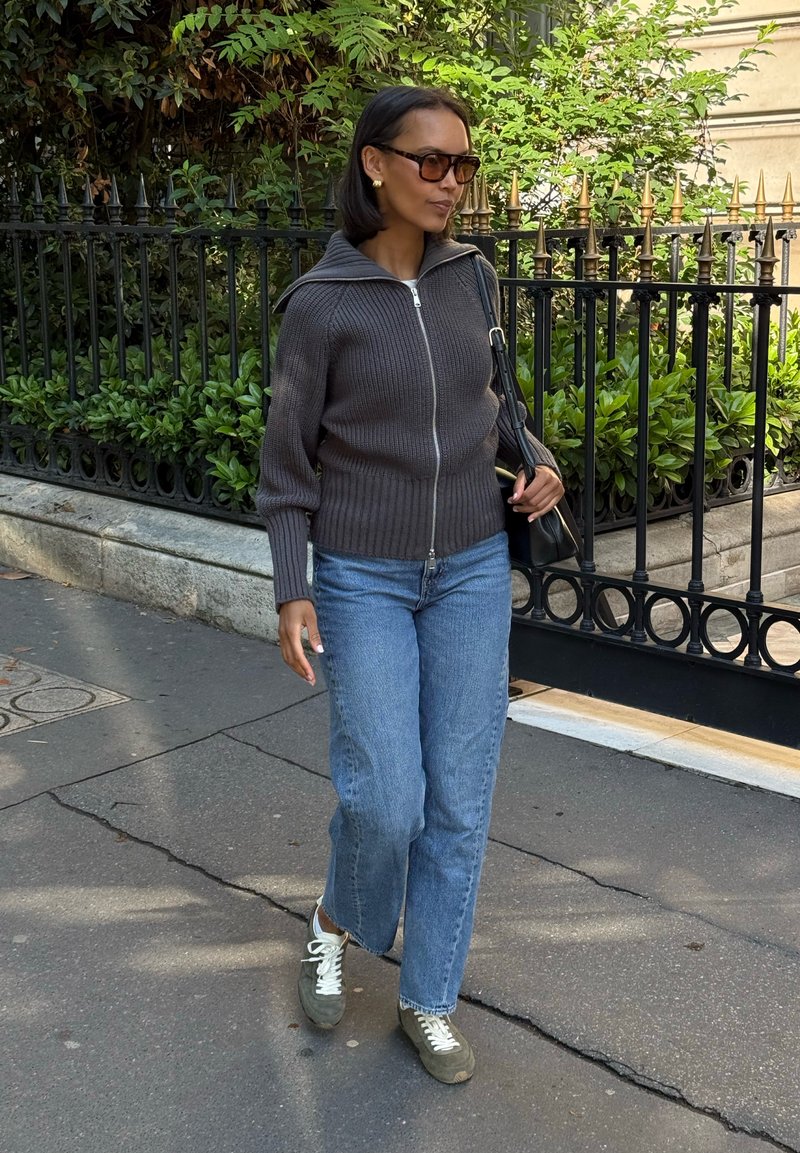 Femme portant des lunettes de soleil, un pull zippé foncé, un jean bleu et des baskets grises marchant sur un trottoir en ville près d'une clôture en fer noir avec de la verdure.