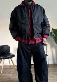 Zwarte bomberjack met een rits aan de voorkant, gedragen over een rood-zwarte geblokte flanellen shirt, gecombineerd met losse zwarte broeken.