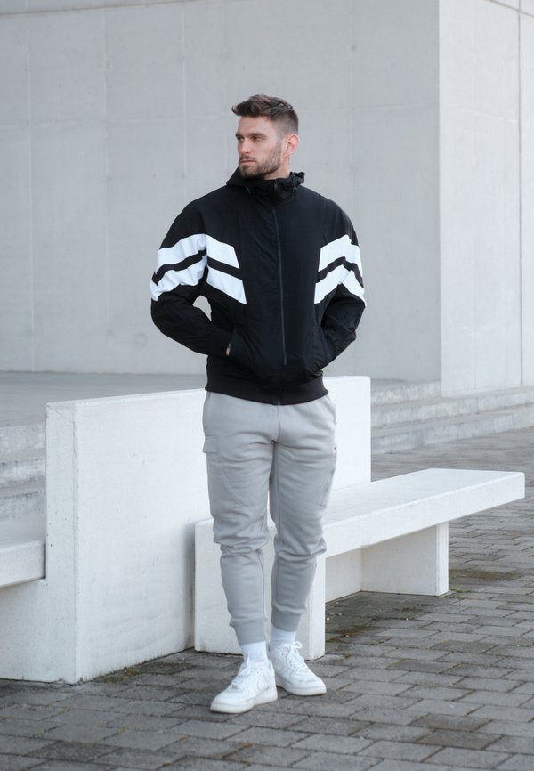 Sort jakke med hvide striber, lynlås foran og hætte; parret med grå sweatpants og hvide sneakers på en belagt overflade.