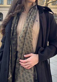 Black coat over a cream cable-knit sweater; an olive green patterned scarf draped around the neck, with a hand resting on the scarf, adorned with silver rings.