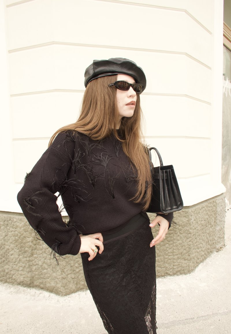 Donna con occhiali da sole neri e basco, maglione nero strutturato e gonna di pizzo, che tiene una borsa nera, in piedi all'angolo di un edificio.