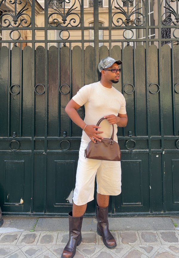 Homme portant une chemise beige, un short blanc, des bottes marron, une casquette camouflage, des lunettes de soleil et tenant un sac à main marron, debout près d'une porte en fer forgé noire.