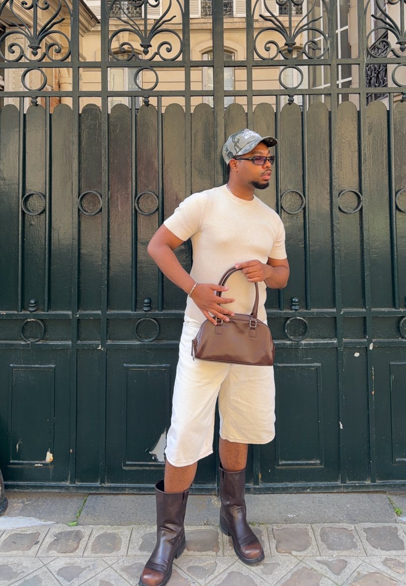 Homme portant une chemise beige, un short blanc, des bottes marron, une casquette camouflage, des lunettes de soleil et tenant un sac à main marron, debout près d'une porte en fer forgé noire.