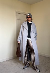 Long grey wool coat, black knit jumper, white collared shirt, striped brown trousers, brown bag, grey beanie, dark sunglasses, silver trainers.
