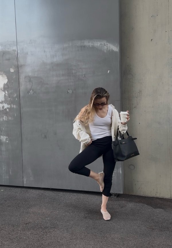 Mujer con camiseta sin mangas blanca y chaqueta, leggings negros, calcetines claros sostiene una taza de café y un bolso negro mientras se equilibra sobre un pie apoyada en una pared gris.