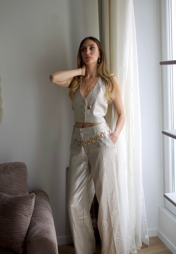 Donna in piedi vicino alla finestra con top crop beige senza maniche e con bottoni e pantaloni a gamba larga con cintura a catena dorata, una mano sul collo e l’altra in tasca.