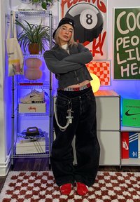 Pantalones vaqueros anchos negros con una cinturilla roja de Diesel, chaqueta gris con cremallera, zapatillas rojas y accesorios plateados, en un entorno interior colorido y moderno.