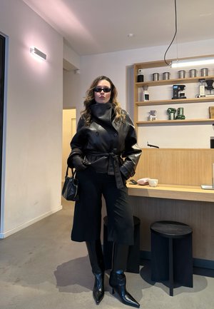 Mujer con chaqueta de cuero negra, guantes, gafas de sol y botas, de pie en una cafetería minimalista con estantes y taburetes de madera.