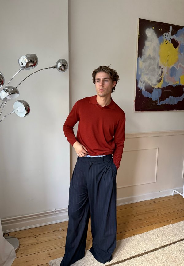 Ung man i röd långärmad skjorta och vida marinblå byxor med smala ränder står inomhus nära en modern silverfärgad golvlampa och en abstrakt väggmålning.