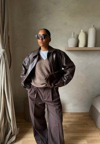 Γυναίκα που φοράει σκούρο καφέ δερμάτινο μπουφάν, καφέ παντελόνι φαρδιάς γραμμής, μπεζ πουλόβερ χωρίς μανίκια, μαύρα γυαλιά ηλίου και κρατάει καφέ τσάντα που ταιριάζει, μέσα σε εσωτερικό χώρο.