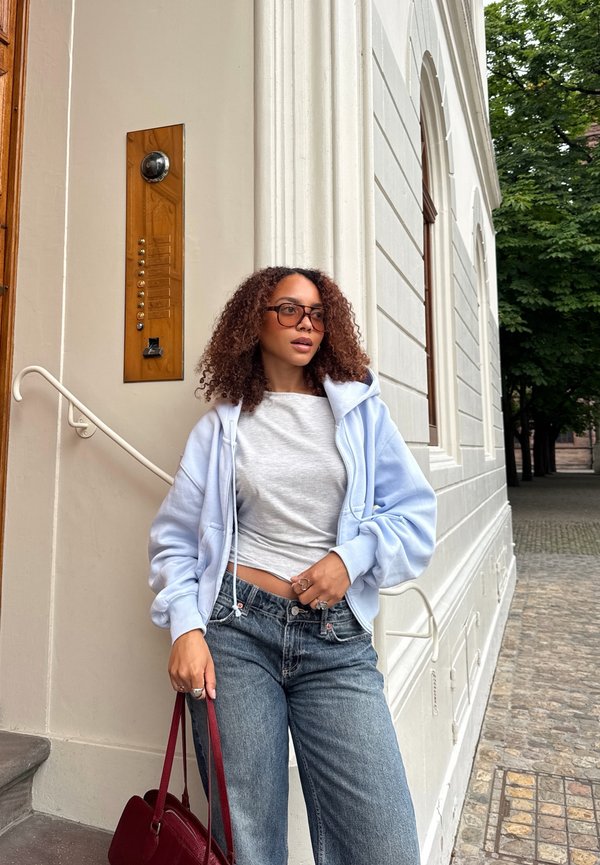 Femme aux cheveux bouclés portant des lunettes, un sweat à capuche bleu clair, un t-shirt gris et un jean, debout près d'un bâtiment blanc tenant un sac rouge dans une rue pavée.