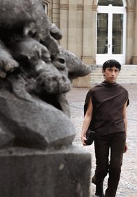 Persoon draagt een donkerbruin mouwloos topje en broek, loopt over kinderkopjes vlakbij een stenen beeldhouwwerk voor de ingang van een gebouw met trappen.