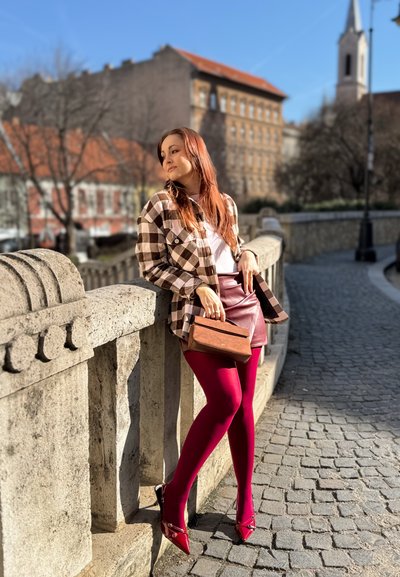 Joven con camisa de cuadros, falda roja y medias rojas apoyada en una barandilla de piedra en una calle empedrada con edificios históricos y una torre de iglesia al fondo.