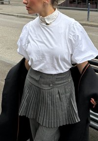 Chemise blanche à manches courtes, jupe plissée grise avec boutons, associée à un manteau noir. Détail notable du col et boucles d'oreilles en forme d'étoile.