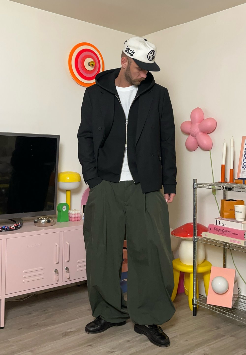Svart dobbeltspent jakke med hette, hvit teksturert skjorte, overdimensjonert grønn bukser og svarte sko; urban setting med fargerik dekor.