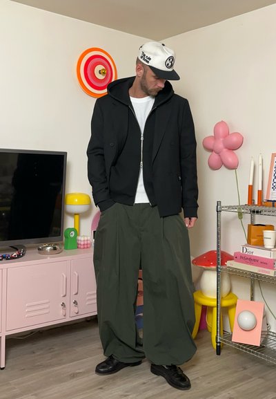 Chaqueta negra de doble botonadura con capucha, camisa blanca texturizada, pantalones verdes de gran tamaño y zapatos negros; entorno urbano con decoración colorida.