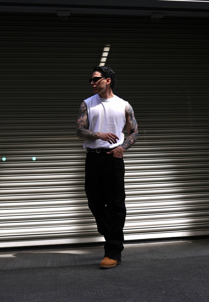Homme tatoué portant une chemise blanche sans manches, un pantalon noir, des lunettes de soleil et des chaussures marron, se tient devant un volet métallique.