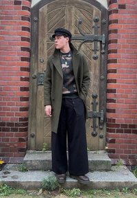 Olivově zelený vlněný kabát přes maskáčový svetr, široké černé kalhoty a hnědé boty. Tmavá čepice, stojící u texturovaných dřevěných dveří.