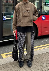 Asmuo, dėvintis rudą striukę, juodą viršutinį drabužį, sluoksniuotas kelnes su languota audinio išorine dalimi, juodus batus, laikantis juodą rankinę šalia raudono autobuso.