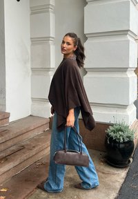Donna che sale scale di pietra, indossa un poncho marrone, jeans blu larghi, scarpe marroni, porta una borsa marrone, accanto a un grande vaso nero con pianta verde.