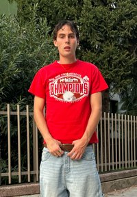 T-shirt rouge en coton avec un motif "Champions" et un casque de football, assorti à un short en denim bleu clair et une ceinture marron.