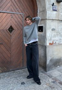 Pull en maille gris à coupe courte, associé à un jean en denim noir ample. Chaussures noires et porte en bois texturée en arrière-plan.