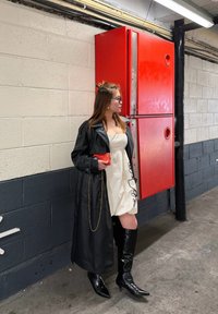 Femme en trench-coat en cuir noir, robe blanche et bottes noires, debout près d'une armoire à tuyaux d'incendie rouge dans un couloir industriel.